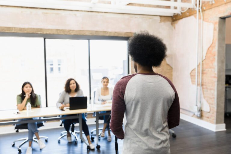 Black actor in a burgundy and gray baseball tee auditions for a role in front of a panel of casting directors.
