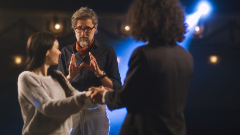An actress and an actor taking notes from their director while rehearsing a scene on a theater stage.