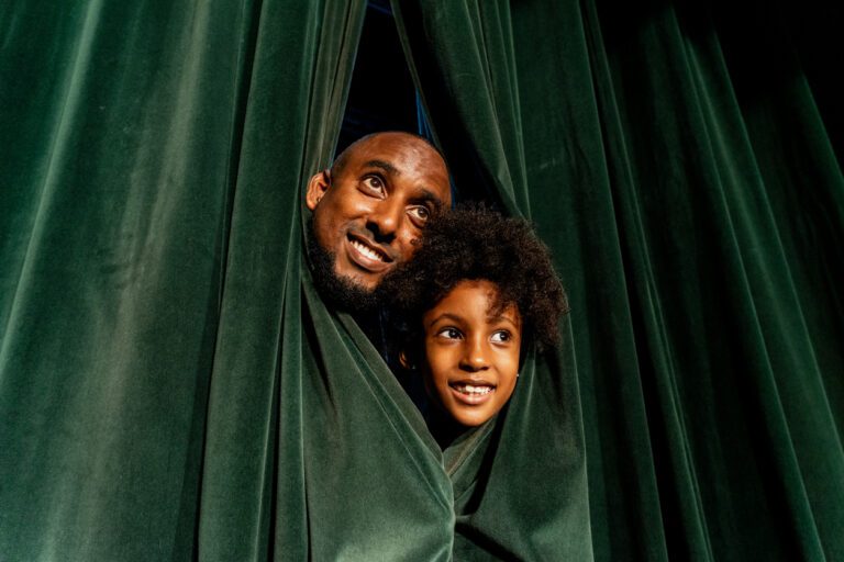 Black actor parent and his son poking their heads out of a green theater curtain.