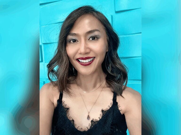 Actor Trevs Begalman smiling in a black dress in front of a blue brick wall.