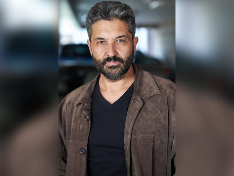 Val Athanassiou in a black t-shirt and a brown leather jacket looking cool in a garage.